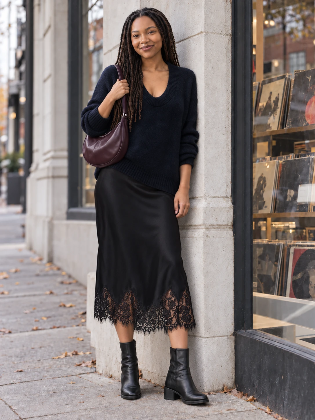 Noelle beside a record-shop window wearing a navy scoop-neck knit, black silk Carolina midi skirt with scalloped black lace hem, black stacked-heel ankle boots and a deep-plum half-moon bag