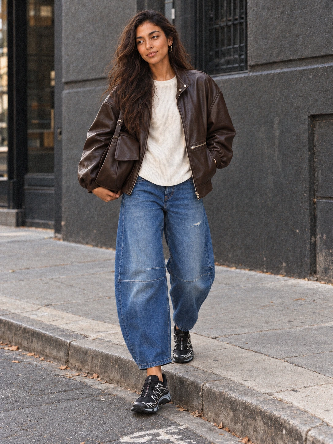 Full-body photograph of Lana stepping off a city curb in a cocoa Veda Harrison leather bomber, warm-ivory thermal, roomy blue barrel jeans, black-and-silver Salomon sneakers and large chocolate flap bag