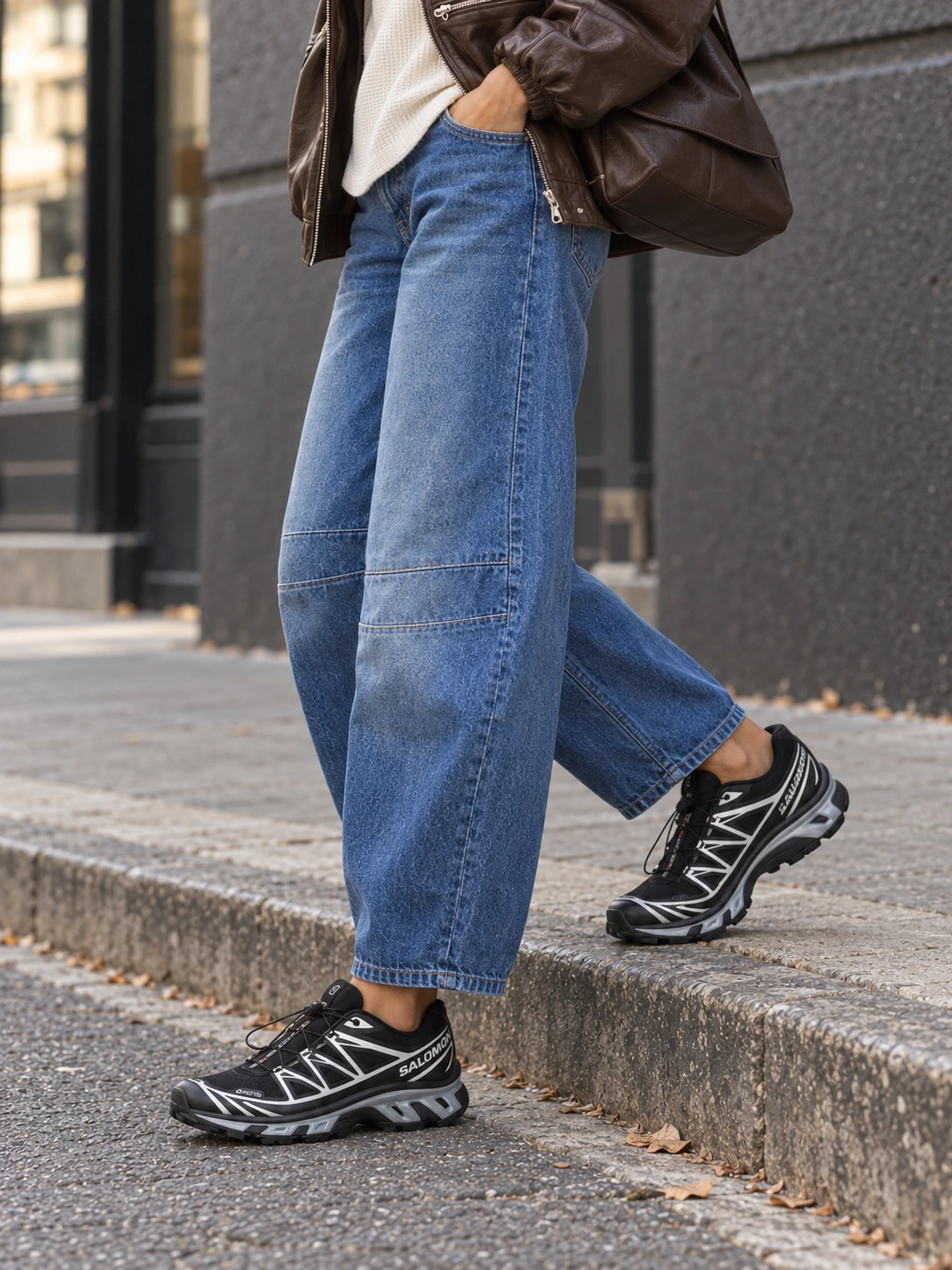 Side-angle detail of both black-and-silver Salomon XT-6 GTX sneakers beside a grey curb, with blue barrel-jean hems and horizontal knee darts visible