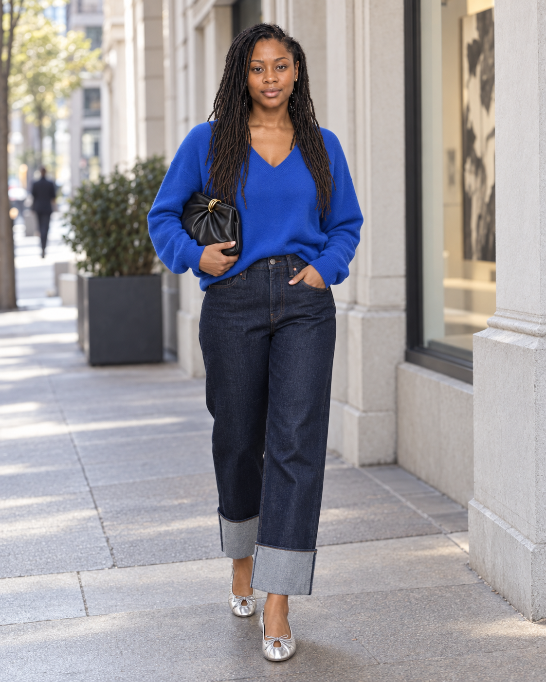 Full-body photograph of curvy Noelle wearing a STAUD cobalt cashmere V-neck, AGOLDE Rinse Big Cuff jeans, silver ALOHAS flats and black Loeffler Randall clutch